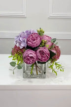 Purple peony rose and hydrangea faux floral centerpiece in a clear glass vase